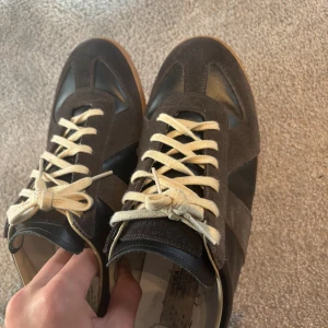 Maison Margiela gats sneakers brun - Maison Margiela gats sneakers i brunt mocka och svart skinn med vita snören och gummisula. Klassisk och stilren modell med rund tå och platt sula. Perfekta för dig som gillar minimalistisk och tidlös design. Skriv vid mer frågor.