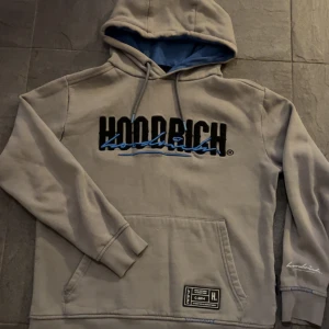 Grå Hoodrich hoodie - Säljer en grå hoodie från Hoodrich i storlek XS. Tröjan har svart och blå logga framtill och baktill, blått foder i huvan och snörning. Tröja är 61cm lång och armarna 57cm lång