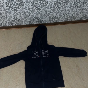Mörkblå hoodie med RM-tryck -  Mörkblå Zip-Hoodie med Koordinat-Design Premium hoodie i djup marinblå färg med exklusiv "R M"-logotyp och koordinater (58°35'48" N, 16°11'35" E). Den stilrena vita texten skapar en modern, minimalistisk look. Hoodien har:  Hel dragkedja för enkel av- och påklädning Mjuk, värmande luva Bekväm passform som passar vardagsbruk Högkvalitativt material som behåller sin form Unisex-design som passar alla stilar Perfekt för den som vill kombinera komfort med ute stil