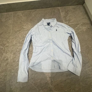 Ljusblå skjorta från Ralph Lauren - Klassisk ljusblå skjorta från Ralph Lauren med lång ärm och knappar framtill. Skjortan har den ikoniska broderade loggan på bröstet och är tillverkad i mjuk bomull. Perfekt för en clean och stilren look.