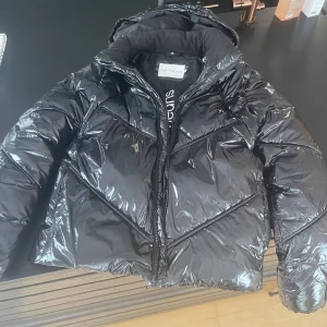 Svart pufferjacka Calvin Klein Jeans - Snygg svart pufferjacka från Calvin Klein Jeans med glansig finish och logga på insidan samt ärmen. Jackan har huva, dragkedja och quiltade detaljer som är väldigt snygg.  Denna jackan är i väldigt bra skick ändast ena jackfickan som inte går att stänga!