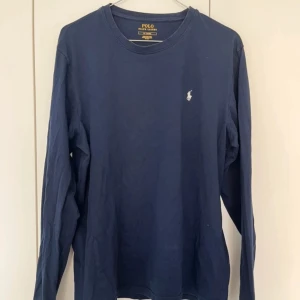 Marinblå långärmad tröja Polo Ralph Lauren - Klassisk marinblå långärmad tröja från Polo Ralph Lauren i mjuk bomull. Tröjan har rund halsringning och den ikoniska broderade loggan i vitt på bröstet. Perfekt basic att ha i garderoben.