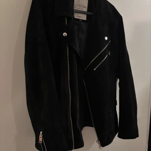 Svart mockajacka från Jack & Jones - Snygg svart mockajacka från Jack & Jones i bikerstil med silvriga dragkedjor och knappar. Jackan har klassisk krage, snedställd dragkedja framtill och dragkedjor vid ärmsluten. Perfekt för dig som vill ha en cool och tidlös look.