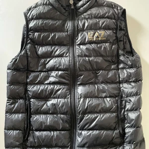 Svart dunväst från Emporio Armani - Snygg svart dunväst från Emporio Armani EA7 med guldfärgad logga på bröstet. Västen har quiltad design, dragkedja framtill och två sidofickor. Perfekt för lager-på-lager och har en modern, sportig look. XL i storlek men passar m-l