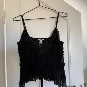Svart volangig linnetopp från New Yorker - Säljer en svart linnetopp från New Yorker i storlek M som är oanvänd och har lappen kvar. Toppen har tunna axelband, volangdetaljer och knyt framtill. Den är i ett lätt, transparent tyg med plisserad struktur och har en croppad passform. Perfekt till fest eller för dig som gillar romantiska och trendiga detaljer!🥰