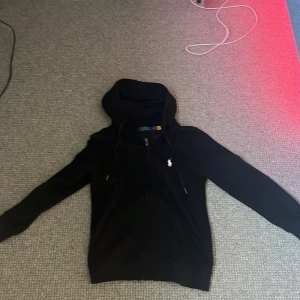 Svart hoodie från Polo Ralph Lauren - Svart hoodie från Polo Ralph Lauren med vit broderad logga på bröstet. Tröjan har dragkedja, huva med snörning och ärmar. Tröjan är i storlek M. Har bild på kvitto! Hör av dig om du har några frågor!🤝