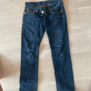 Mörkblå jeansbyxor True Religion - Snygga mörkblå jeansbyxor från True Religion med klassiska kontrastsömmar och ikoniska bakfickor med lock och knapp. Byxorna har låg midja och raka ben, tillverkade i slitstarkt bomullsjeansmaterial. Om du har frågor, tveka inte🙋‍♂️🤝🏽