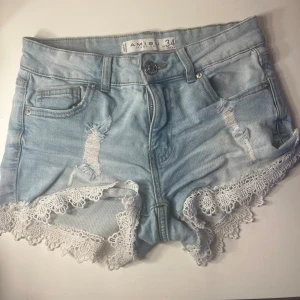 Ljusblå jeansshorts med spets från Amisu - Snygga ljusblå jeansshorts från Amisu med slitna detaljer och vit spetskant längst ner. Modellen är högmidjad med klassiska fem fickor och knappstängning fram. Perfekt för dig som gillar en feminin och trendig look på sommaren.