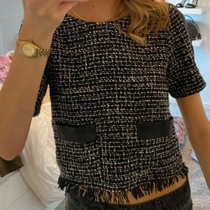 Svartvit rutig blus med fransar - Säljer en svartvit rutig blus med korta ärmar och coola fransdetaljer längst ner. Blusen har två svarta dekorativa fickor framtill och svarta paneler på ärmarna. Materialet är grovt vävt och ger en lyxig känsla, perfekt för dig som gillar statement-plagg. Ända defekten är att de är lite slitet på ”skin” partierna på axlarna, 