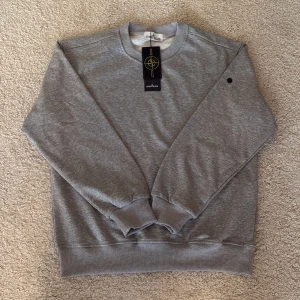 Grå sweatshirt från Stone Island - Grå sweatshirt från Stone Island med klassisk rund hals och ribbade muddar vid ärmslut och nederkant. Tröjan har två svarta knappar på ena ärmen för att fästa den ikoniska Stone Island-patchen. Mjuk bomullskvalitet och stilren design. Storlek S/M.