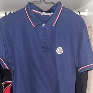 Marinblå Moncler pikétröja M - Snygg marinblå pikétröja från Moncler med klassisk krage och rödvit rand på krage och ärmslut. Tröjan har korta ärmar, knappar framtill och Moncler-logga på bröstet. Perfekt för dig som gillar stilrena plagg.