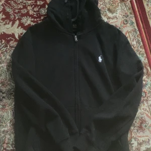 Polo Ralph Lauren hoodie  - Svart hoodie från Polo Ralph Lauren i custom fit med dragkedja framtill och klassisk huva. Tröjan har två fickor på magen och den ikoniska vita loggan broderad på bröstet. Perfekt för en chill och stilren look.