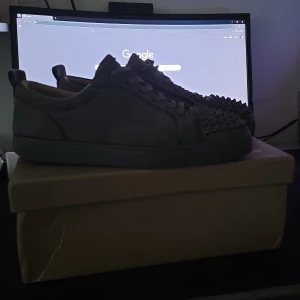 Christian Louboutin sneakers med nitar - Säljer ett par mörkgrå sneakers från Christian Louboutin i mocka med ikoniska röda sulor och coola nitar framtill. Skorna har snörning och en klassisk låg siluett. Perfekta för dig som vill sticka ut med exklusiv streetstyle. Pris kan diskuteras