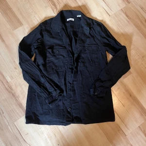 Svart overshirt från Jack & Jones - Svart overshirt från Jack & Jones i storlek S. Skjortan har klassisk krage, knäppning framtill och två bröstfickor med lock. Tillverkad i ett lätt bomullstyg, perfekt att slänga över en t-shirt för en chill look.