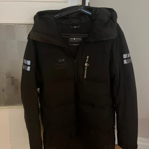 Svart pufferjacka från Sail Racing - Svart pufferjacka från Sail Racing med huva och logga på bröstet, ärmarna och huvan. Jackan har dragkedja framtill och är vadderad för extra värme. Perfekt för kalla dagar och har en clean, sportig look. Jackan är i storlek small men passar mig som är 187cm. 