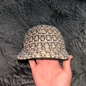 Salvatore Ferragamo beige keps - Snygg keps från Salvatore Ferragamo i beige och svart med ikoniskt mönster över hela kepsen. Justerbar rem baktill med metallspänne och läderdetalj med logga. Tillverkad i bomullstyg med svart innerfoder. Perfekt accessoar för en trendig look.