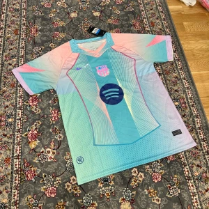 FC Barcelona x Nike pastell fotbollströja M - Säljer en FC Barcelona fotbollströja från Nike i pastellfärger med coolt grafiskt mönster och Spotify-logga på bröstet. Tröjan är kortärmad, tillverkad i lätt Dri-FIT-material som andas. Färgerna går i blått, rosa och ljus turkos med vita detaljer.