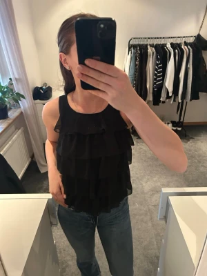 Svart volangig linnetopp - Säljer en svart ärmlös linnetopp med flera lager volanger framtill. Toppen har rund halsringning, baktil är det bara slätt tyg. Det är barnstorlek 152 men passar mig som är xs 