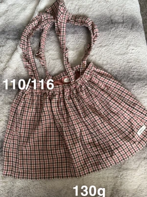 Rutig kjol med hängslen från Newbie - Supersöt rutig kjol från Newbie med justerbara hängslen och knappar. Kjolen har en klassisk rosa, vit och brun färgkombination. Perfekt för dig som gillar en lekfull och trendig stil.🌸 Oanvänd. Storlek 110/116