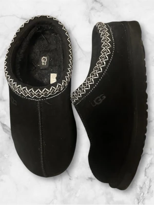 UGG skor - UGG tofflor i svart mocka med fluffigt foder och dekorativt vitt zigzag-mönster runt öppningen. Platt sula i gummi och rund tå. Perfekta för att hålla fötterna varma under kyliga dagar. Klassisk UGG-logga på sidan.