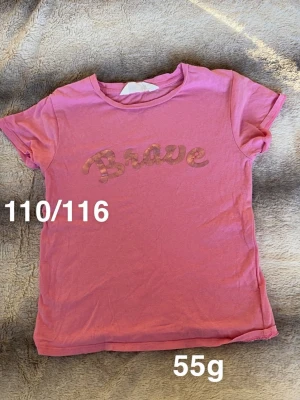 Rosa t-shirt med Brave-tryck - Söt rosa t-shirt med texten 'Brave' i glittrigt tryck på bröstet. Klassisk rund halsringning och korta ärmar. Perfekt för dig som gillar färg och vill ha en enkel men cool look.🌸 Fint använt skick. Storlek 110/116 men mer som 110