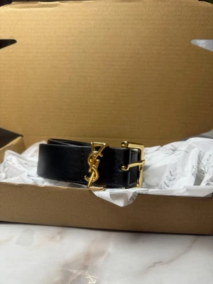 Svart YSL bälte med gulddetaljer - Säljer ett svart bälte från YSL i slätt skinn med ikoniskt YSL-logo i guld framtill. Bältet har en fyrkantig guldfärgad metallspänne som ger en lyxig touch. Perfekt accessoar för att lyfta vilken outfit som helst.