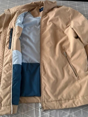 Beige vindjacka från Gant - Snygg beige vindjacka från Gant med minimalistisk design och diskret logga på bröstet. Jackan har två snedställda fickor med dragkedja, hög krage och hel dragkedja framtill. Insidan har kontrastfärger i blått och ljusblått. Perfekt för vår och höst.
