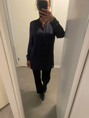 Mörkblå silkeslen skjorta Bik Bok XS - Mörkblå skjorta från Bik Bok i silkesliknande material men i själva verket konstmaterial/polyester. Långärmad modell med guldknappar framtill, på axlar och vid ärmslut. Skjortan har en rak passform och snygg v-ringning utan krage. Perfekt för en clean och stilren look. Superfin, elegant och kvinnlig. Passar även small