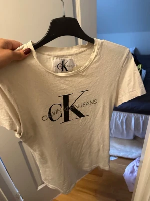 Vit t-shirt från Calvin Klein Jeans - Säljer en klassisk vit t-shirt från Calvin Klein Jeans med stor svart CK-logga framtill. T-shirten har rund halsringning och korta ärmar. Tillverkad i mjuk bomull för en skön känsla och enkel stil.