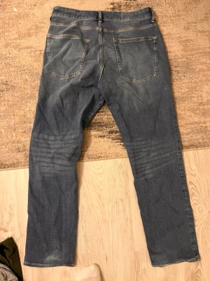 Blå raka jeans med klassisk tvätt - Säljer ett par blå jeans med rak passform och klassisk femficksdesign. Jeansen har normal midja och subtila slitningar framtill för en avslappnad look. Tillverkade i tåligt denimtyg som passar till det mesta. Perfekta för dig som gillar en enkel och tidlös stil.