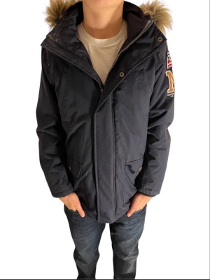Marinblå parkas från Hampton Republic - Säljer en marinblå parkas från Hampton Republic med snygg fuskpäls på huvan och coola patches på ärmen. Jackan har dragkedja, knappar och praktiska fickor framtill. Perfekt för kalla dagar och riktigt skön att bära.
