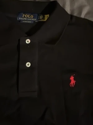 Svart piké från Polo Ralph Lauren - Klassisk svart pikétröja från Polo Ralph Lauren med röd broderad logga på bröstet och vita knappar. Tröjan har korta ärmar och normal passform, perfekt till jeans eller shorts. Materialet är mjuk bomull som känns skön mot huden.