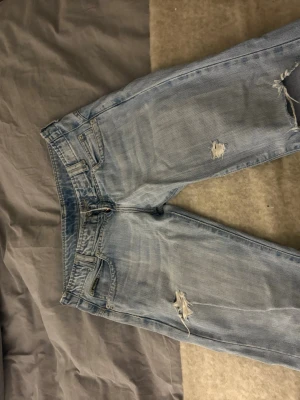 Ljusblå slitna jeans med broderade fickor - Säljer ett par ljusblå jeans med slitningar och hål på benen för en avslappnad vibe. Jeansen har raka ben och snygga broderade detaljer på bakfickorna. Klassisk femficksmodell i mjukt denimtyg, perfekta till sneakers eller boots.