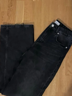Svarta raka jeans från Zara - Säljer ett par svarta jeans från Zara med rak passform och rå kant nedtill. Klassisk femficksmodell med silverfärgade nitar och knapp. Jeansen har en enkel, stilren look och är tillverkade i ett robust denimtyg.