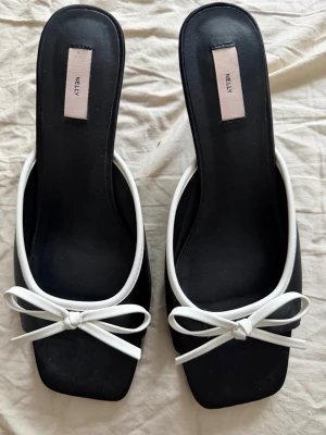Svarta klackade sandaler från Nelly - Säljer ett par stilrena svarta sandaler från Nelly med vit kant och rosett framtill. Skorna har fyrkantig tå och smal hög klack. Materialet är syntet och designen är enkel men trendig, perfekt till sommarens outfits.