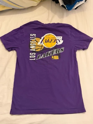 Lakers t-shirt lila från New Era - Säljer en lila Los Angeles Lakers t-shirt från New Era i storlek S. T-shirten har tryck med Lakers-logga och basketboll både fram och bak samt NBA-detalj. Klassisk passform med korta ärmar och rund hals. Materialet är mjuk bomull.