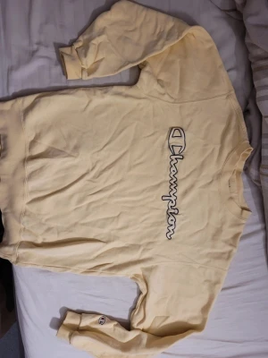 Gul sweatshirt från Champion - Säljer en ljusgul sweatshirt från Champion med stor broderad logga på bröstet och liten logga på ärmen. Tröjan har rund hals och långa ärmar, perfekt för dig som gillar en avslappnad och sportig stil.
