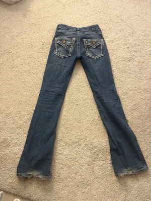 Blå bootcut jeans med kontrastsömmar - Säljer ett par blå jeans med bootcut passform och tydliga kontrastsömmar på bakfickorna. Jeansen har klassisk femficksdesign, knapp och dragkedja fram samt slitna detaljer vid bensluten för en avslappnad look. 