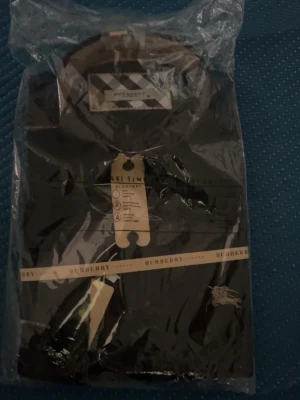 Svart skjorta från Burberry - Snygg svart skjorta från Burberry med klassisk krage och diskret broderad logga på bröstet. Skjortan har knappar framtill och detaljer i Burberrys ikoniska rutmönster på insidan av kragen. Perfekt för dig som gillar stilrena plagg med exklusiv känsla. Storlek M/S