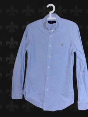 Blå skjorta från Ralph Lauren - Klassisk ljusblå Ralph Lauren Oxford-skjorta i Slim Fit. Tidlös modell med button-down-krage och broderad logga på bröstet. 	•	Storlek: S 	•	Färg: Ljusblå 	•	Skick: Inga tecken på användning! 	•	Material: Bomull 	•	Passar både till vardag och mer uppklätt  Rök- och djurfritt hem. Skickas snabbt 📦