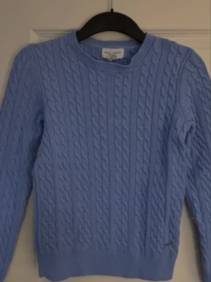 Blå kabelstickad tröja Holly & Whyte - Säljer en ljusblå kabelstickad tröja från Holly & Whyte. Tröjan har rund halsringning, ribbade muddar och lång ärm. Perfekt för lager på lager under höst och vinter. 