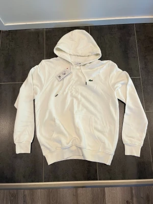 Lacoste Hoodie - Säljer denna stilrena, nya och riktigt snygga vita Lacoste hoodien, passar perfekt till allt, dock är den liten i storleken, står XL men passar (M-L), skriv gärna vid intresse och för mer information!