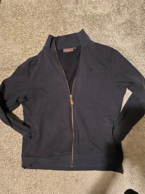 Mörkblå zip-kofta från Morris - Säljer en mörkblå kofta från Morris (i stl M men sitter liten i stl så passar mer som XS elr S) med hel dragkedja och diskret logga på bröstet. hög krage och är tillverkad i mjuk bomull. Har andvänt den mycket få gånger då jag råka krympa den i tvätten. Nypriset ligger på runt 1000kr men ja säljer den för ändast 250kr och inga defekter. Vid frågor kom dm🤩🤗