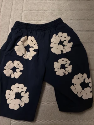 Blå shorts från Kapital med blommönster - Unika marinblå shorts från Kapital med vita blommiga cirkelmönster över hela plagget. Elastisk midja för skön passform och tillverkade i mjuk bomull. Perfekta för dig som vill sticka ut med en cool och kreativ stil. en liten defekt men inget som märks och gör säkert bort med lite starkare tvättmedel 