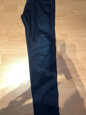 Replay mörkblå jeansbyxor herr - Säljer ett par mörkblå jeans från Replay i klassisk femficksmodell. Jeansen har normal passform och raka ben, tillverkade i slitstarkt bomullsmaterial. Snygga detaljer med Replay-logga på fickan och metallknapp i midjan.  Aldrig använt 