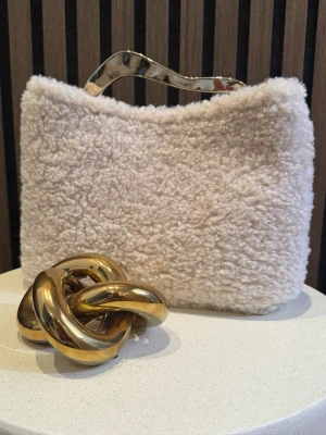 River Island Handväska  - Supersnygg beige handväska i fluffigt syntetmaterial med unikt vågigt metallhandtag och vita detaljer. Perfekt accessoar för dig som vill sticka ut och addera lite lyxig känsla till din outfit. Väskan har en mjuk, fårullsinspirerad look och känns riktigt trendig.