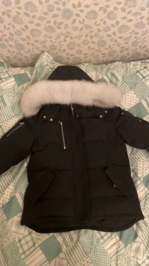 Svart Moose Knuckles dunjacka med päls - Säljer en svart dunjacka från Moose Knuckles med stor vit päls runt huvan. Jackan har flera fickor med dragkedja, snygga metalldetaljer och logga på ärmen. Perfekt för kalla dagar och riktigt stilren look. Använt max 1 gång, ångrar köpet, kan gärna ta emot byten om det finns och pris går ALLTID att diskutera!