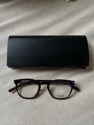 Saint Lauren glasögonbåge - Saint Laurent båge att sätta styrkeglas i  Saint Laurent fodral med tillhörande putsduk ingår  Modell: SL30 SLIM Färg: 003 (mörkbrun/melerad) Strl: 49-23 150  Nypris: ca 3000:-   Glasögonen är helt oanvända, i originalskick och ej justerade.  Köptes till sambo, men där det aldrig blev av att glasa dem, så de har alltså bara legat oanvända i fodralet.  Bågen är unisex. Hänger ofta på herr sektionen, men fungerar lika bra till kvinnor, dock är riskerar den att bli lite bred över näsan och hamna lå