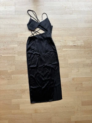 Svart långklänning med cut out och smala band - Säljer en svart långklänning med smala axelband och trendiga cut out-detaljer i midjan. Klänningen har en figurnära passform och är tillverkad i ett glansigt, mjukt material som ger en lyxig känsla. Perfekt för dig som vill sticka ut med en modern och stilren look.