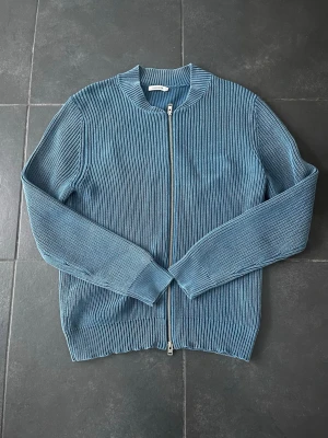 Blå ribbad kofta från J.Lindeberg - Säljer en snygg blå ribbad kofta från J.Lindeberg i storlek M. Koftan har dragkedja framtill, rund halsringning och är långärmad. Materialet är stickat och ger en schysst struktur. Perfekt för dig som gillar stilrena och bekväma plagg.